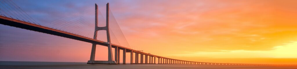A modern bridge spans a large waterway and curves into the distance with a brilliant sunset in the background
